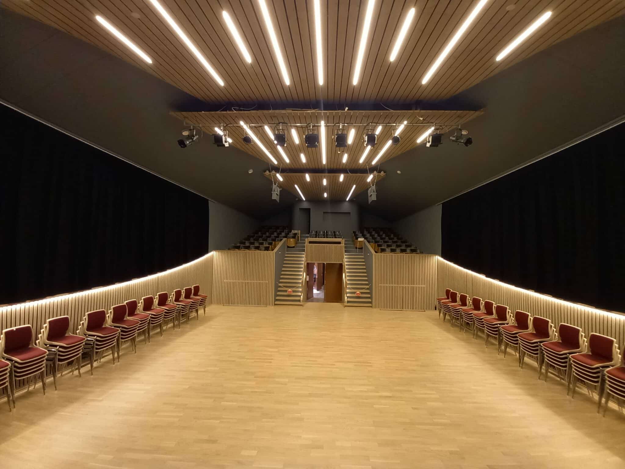 Et tomt, moderne flerbrukshus eller teatersal med lyst tregulv. Røde stoler er stablet langs begge sideveggene. I bakgrunnen fører to trapper opp til et galleri med faste sitteplasser. Taket har dekorative trelameller og integrert lineær belysning.