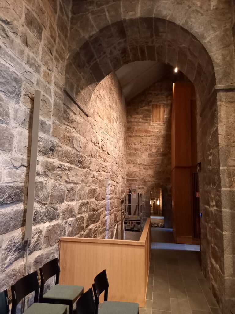 En smal gang eller sideareal i en kirke med vegger av grovhugget stein. En moderne lift står parkert i bakgrunnen, og en lav skillevegg i lyst treverk rammer inn området i forgrunnen.