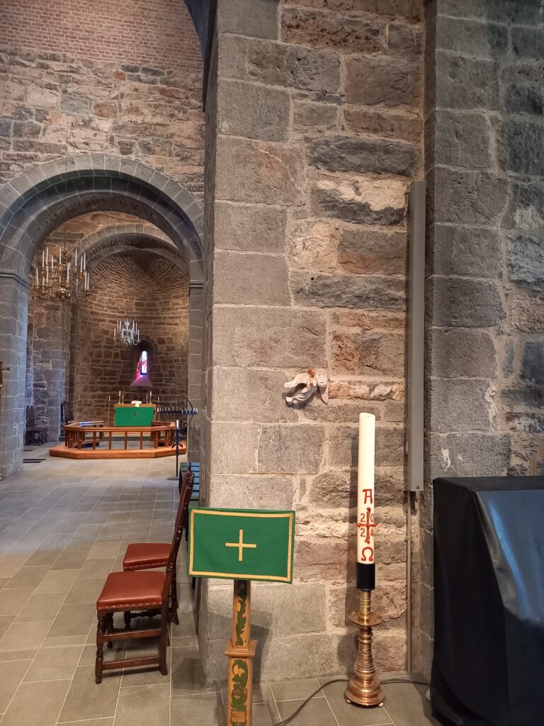 Nærbilde av en massiv steinpilar i en kirke. På pilaren er det montert en liten englefigur i relieff og et høyt påskelys står på en stake foran. I bakgrunnen ses koret med et grønt alterklede og en rund alterring.