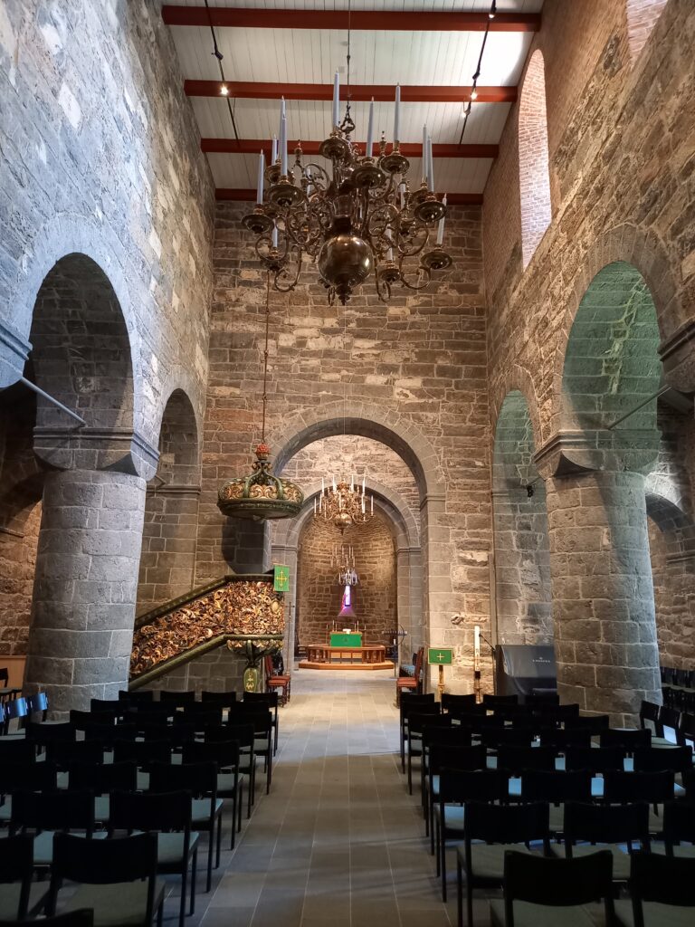 Et perspektivbilde nedover midtgangen i en steinkirke mot alteret. Svarte stoler er stilt opp i rader. To store messingkroner henger fra taket, og den detaljerte prekestolen er synlig til venstre.