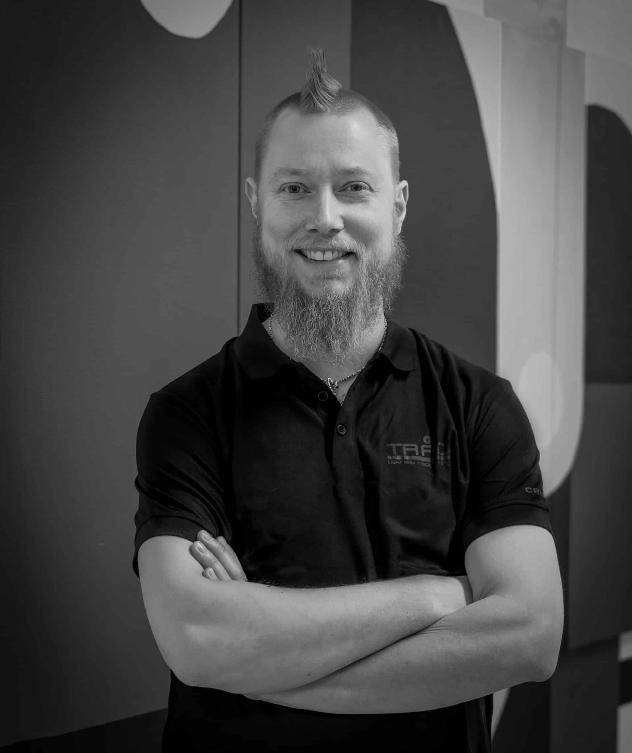 Svart-hvitt portrett av en smilende mann med skjegg og en liten hanekam-frisyre. Han har armene i kors og er iført en mørk polo-skjorte med en diskret logo.