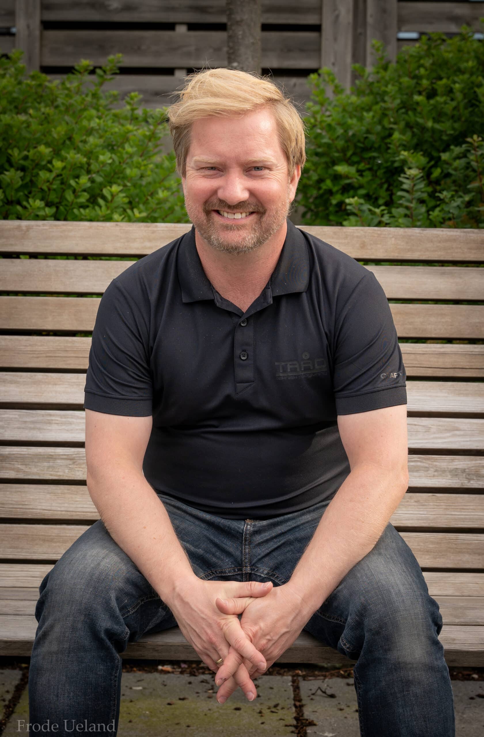 Portrett av en smilende mann med lyst, litt rufsete hår og skjegg som sitter på en trebenk utendørs. Han er iført en svart polo-skjorte og har hendene foldet i fanget.