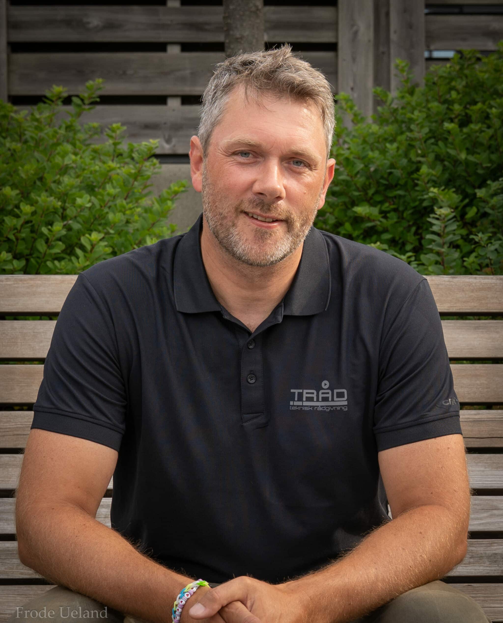 Portrett av en mann med kort, gråsprengt hår og skjegg som ser direkte inn i kameraet med et vennlig uttrykk. Han sitter utendørs foran grønne busker og bærer en svart polo-skjorte med "TRÅD"-logo.