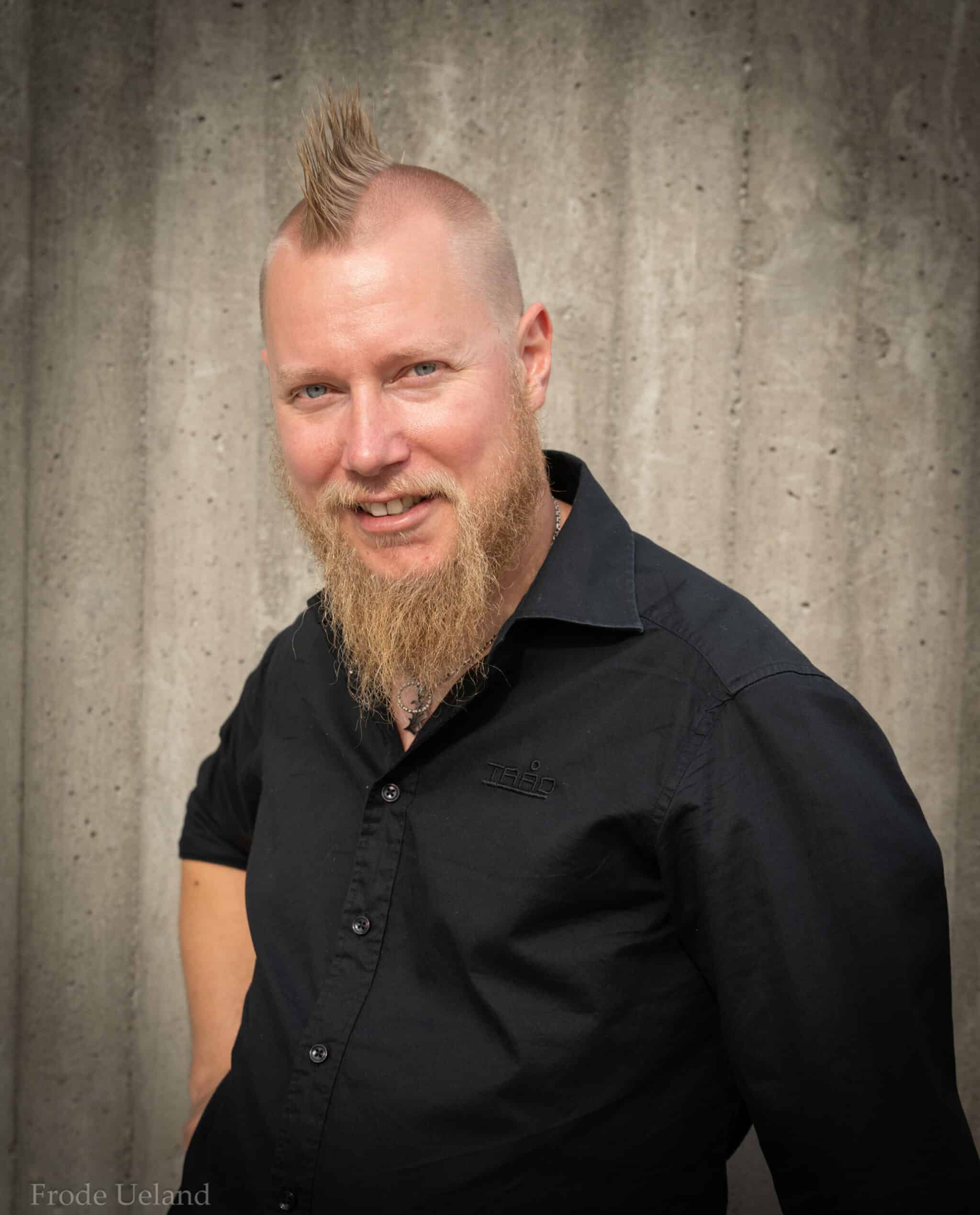 Portrett av en smilende mann med lyst skjegg og en markant hanekam-frisyre. Han er iført en svart skjorte med en diskret logo og står foran en grå betongvegg.