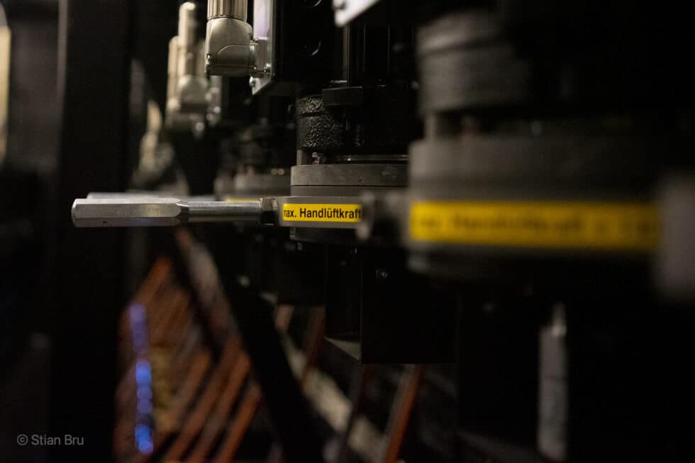 Detaljbilde av mekanisk utstyr, sannsynligvis en del av et scenetrekk-system. En gul etikett med teksten "max. Handlüftkraft" er synlig på en av de svarte metallkomponentene.