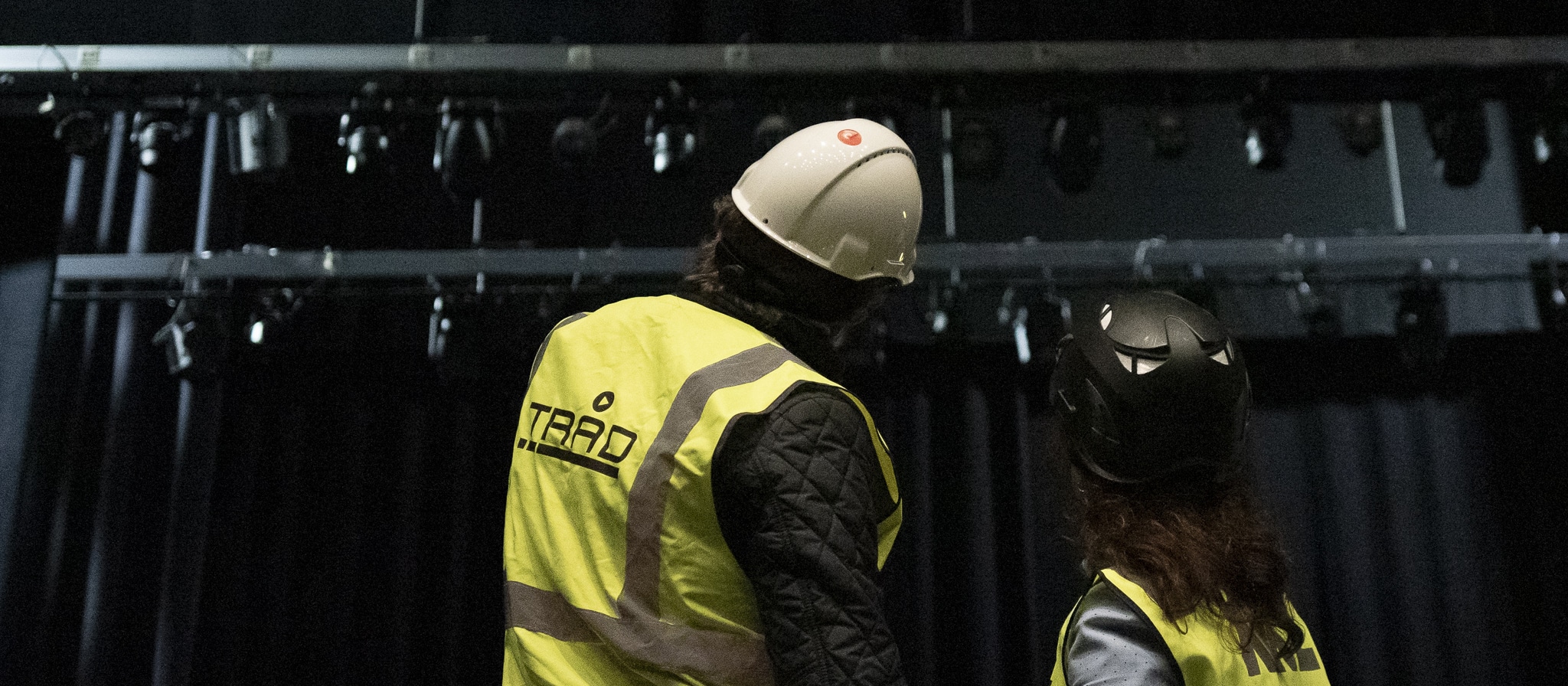 To personer sett bakfra, begge iført gule refleksvester med "TRAD"-logoen. Den ene har hvit vernehjelm og den andre svart klatrehjelm. De ser opp mot en rigg med teaterlyskastere i et mørkt scenerom.