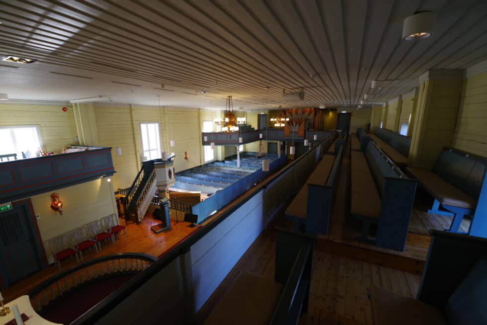 Interiøret i en historisk steinkirke (Stavanger domkirke). En rikt utskåret og fargerik prekestol i barokkstil dominerer venstre side. En stor messingkrone henger fra taket i midtgangen som fører mot alteret i bakgrunnen.
