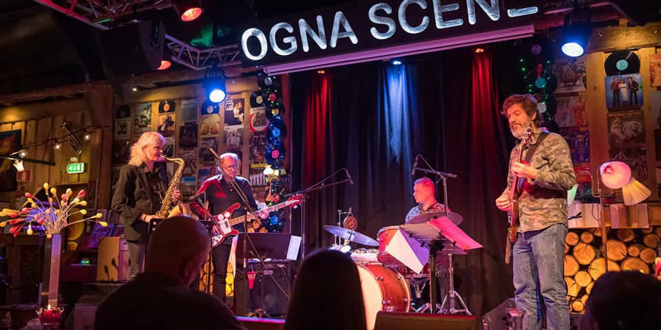Et live-band spiller på en intim scene med navnet "OGNA SCENE" i lysskilt over scenen. Bandet består av en saksofonist, bassist, trommeslager og gitarist. Veggen bak er dekorert med LP-plater, og belysningen er varm med blå og røde fargetoner.