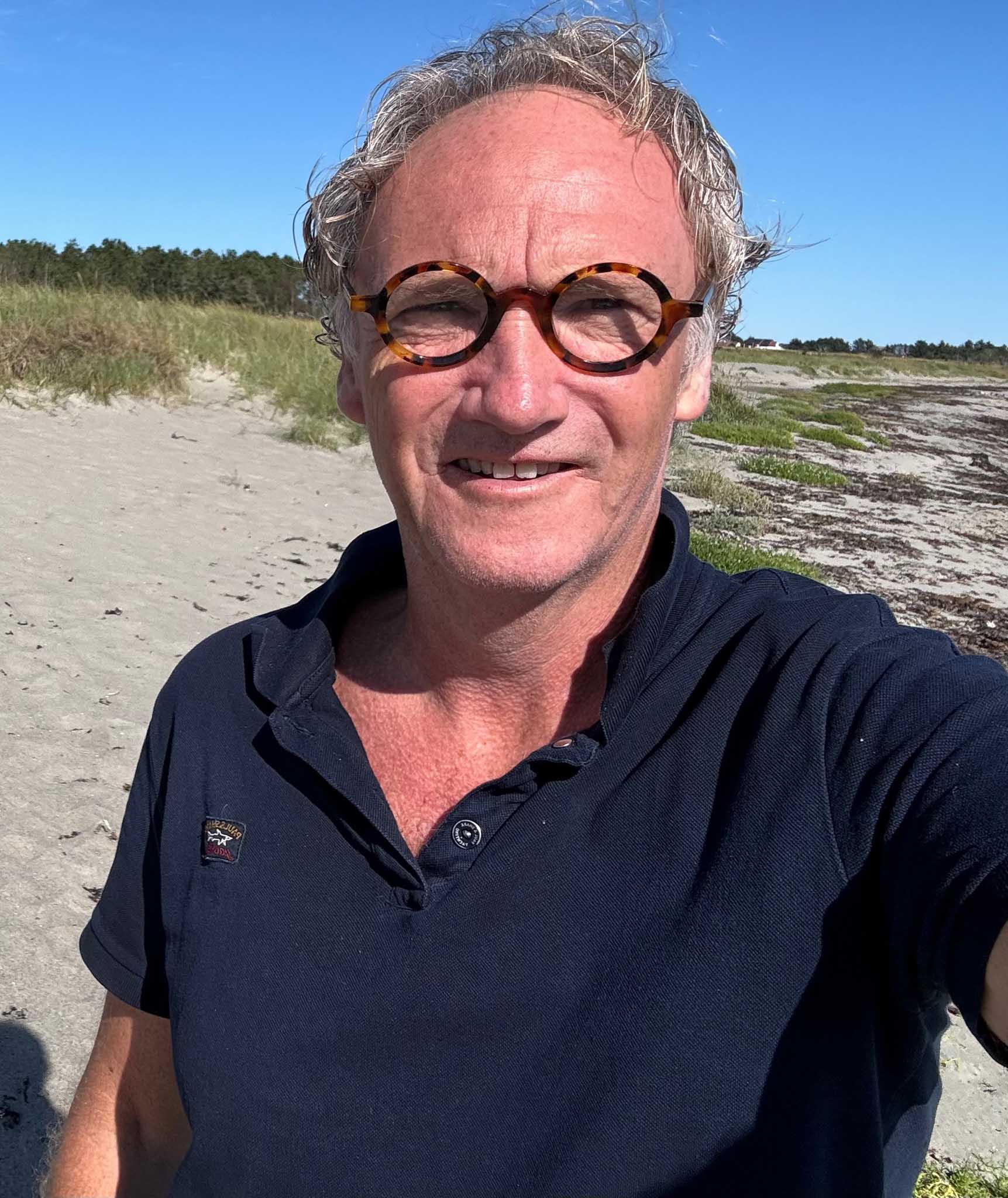 Portrett av en mann med briller og mørk blå polo-skjorte som står utendørs på en strand. Bak ham er sanddyner og en klar, blå himmel.